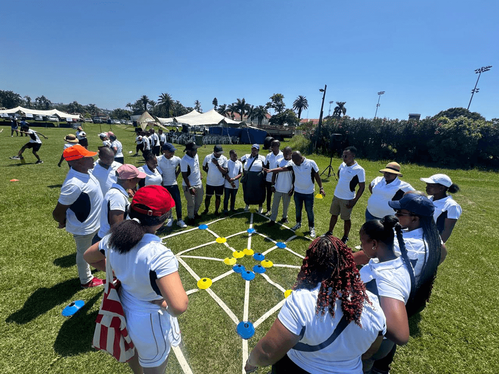Corporate teambuilding exercise on a sunny sports field, with colleagues in matching white shirts gathered in a circle around a strategy game using colorful cones and ropes.